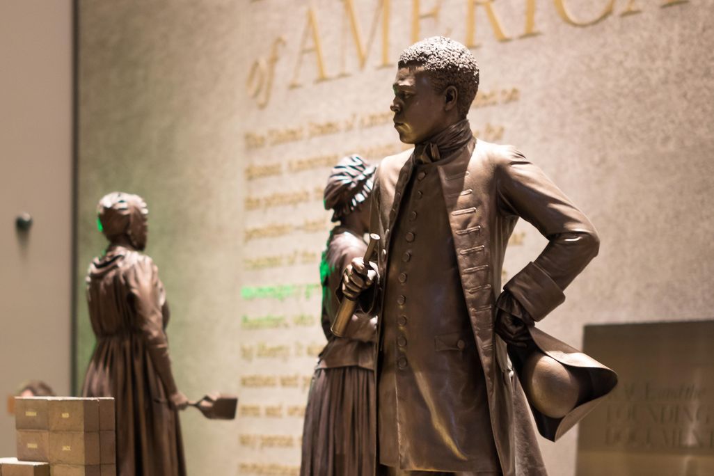 A bronze colored statue of Bannecker, a Black man wearing a suit jacket and high boots, stands in a museum exhibition