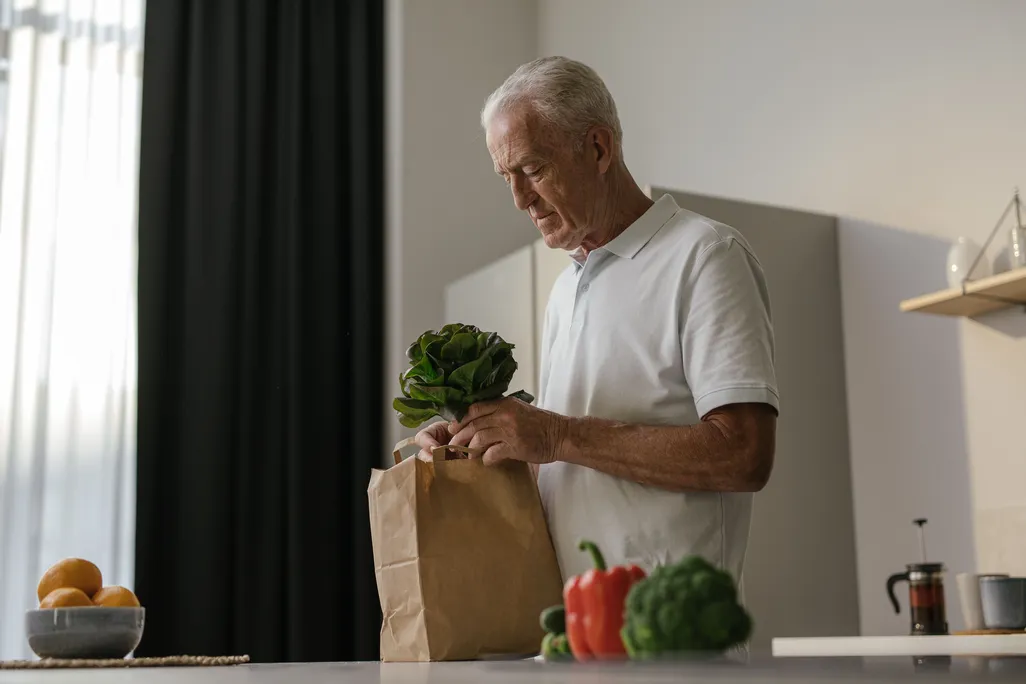 An older man putting away groceries that are mainly vegetables