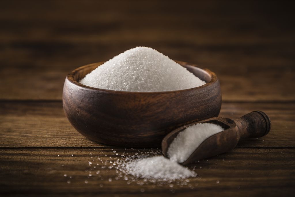 White sugar in a brown wooden bowl with a serving scoop in front with sugar pouring out