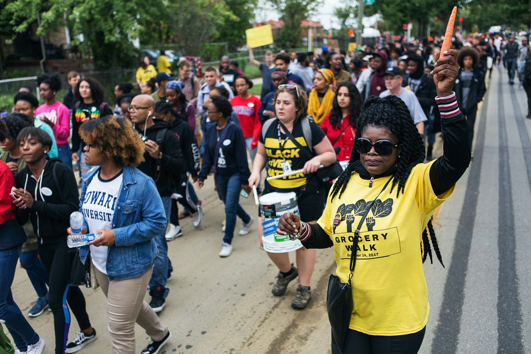 A Black woman wearing a yellow Grocery Walk 2017 t-shirt brandishes a carrot in the air, surrounded by a diverse crowd of other protesters.