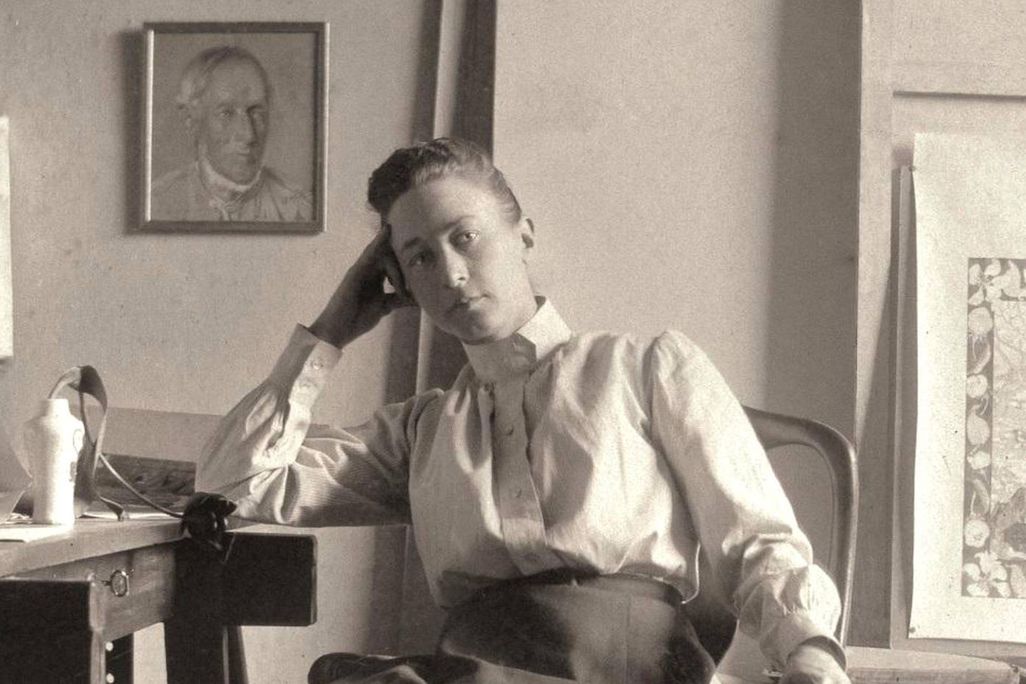 Hilma af Klint, a white woman in a white shirt and a long black skirt, leans her hand on one elbow and sits at a desk in her studio