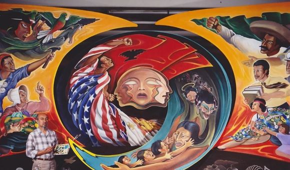 Leo Tanguma stands in front of a large mural-in-progress that features mythical figures, revolutionaries, laborers, and other figures of Mexican and Chicano history.