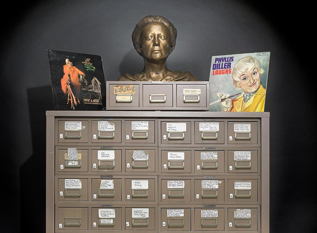 Partial photo of Phyllis Diller's gag file at the Smithsonian with a small three drawer file on top along with two of her record albums and a bronze bust of her head