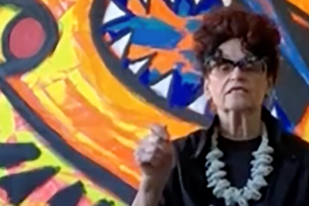 Detail of color screenshot of Judith Bernstein in front of one of her paintings wearing a black blouse and light-colored, chunky necklace holding her hand up in a fist as if making a point in conversation.