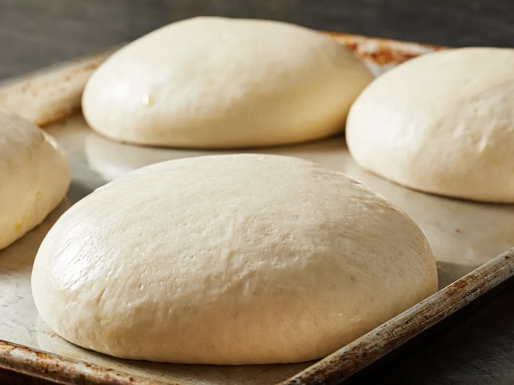 Four balls of pizza dough rising on a baking sheet