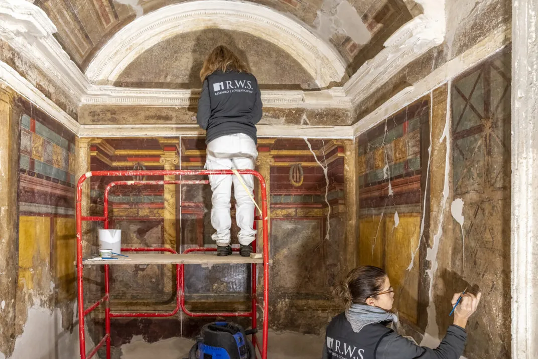 A person standing on scaffolding with another person working on a wall nearby