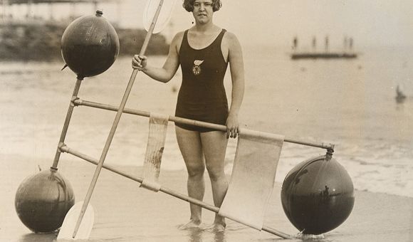 A black and white gelatin silver print of Gertrude Ederle standing on the shore with a 1920s flotation device.
