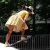 A woman in a yellow dress and long braids skateboarding on a half-pipe