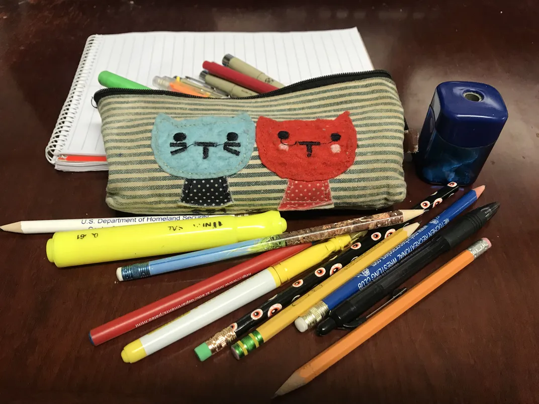 A notebook, pencil holder, and pencil sharpener along with several pencils, pens, and highlighters on a wooden table.