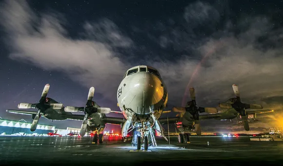 A massive aircraft with four propellers sits on a wet tarmac reflecting the bright red and white lights of the surrounding air base. The night sky above is cloudy, though some bright stars are visible.