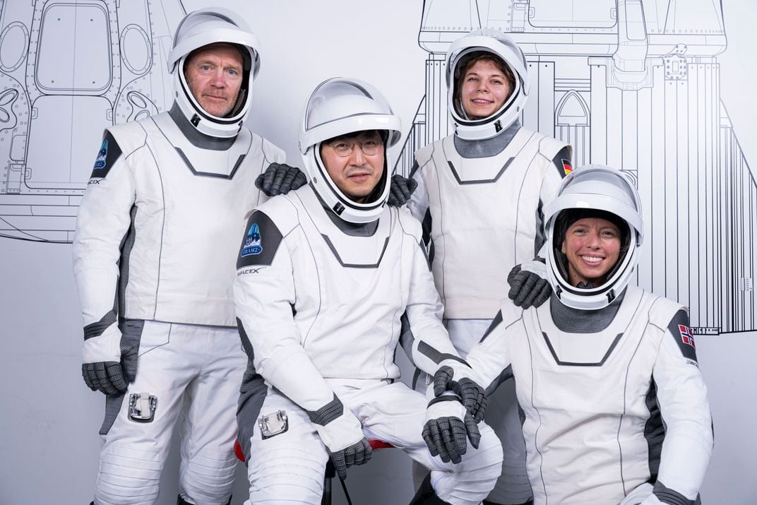 four astronauts in white suits pose in front of a white wall with sketches of a rocket and spacecraft
