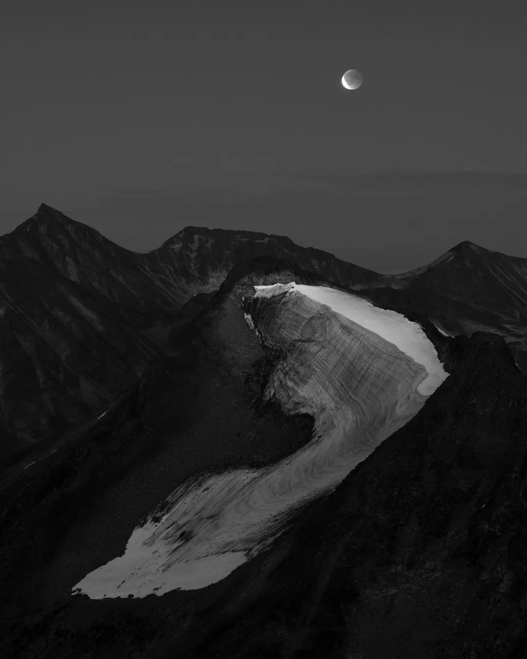 in black and white, the full moon in partial shadow sits above mountains with a glacier