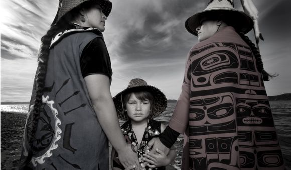 Darkfeather Ancheta, Eckos Chartraw-Ancheta, and Bibiana Ancheta, Tulalip, Washington. Darkfeather, is pictured with her sister Bibiana and nephew Eckos at the edge of Tulalip Bay. They are wearing traditional regalia prepared for their annual Canoe Journey. Every year, upwards of 100 U.S. tribes, Canadian First Nations and New Zealand canoe families will make “The Journey” by pulling their canoes to a host destination Tribe. Canoe families pull for weeks, and upon landing participate in several days and nights of “protocol”, a celebration sharing traditional knowledge, ancestral songs, and sacred dances. Photo by Matika Wilbur for Project 562. Courtesy of the Artist. 
