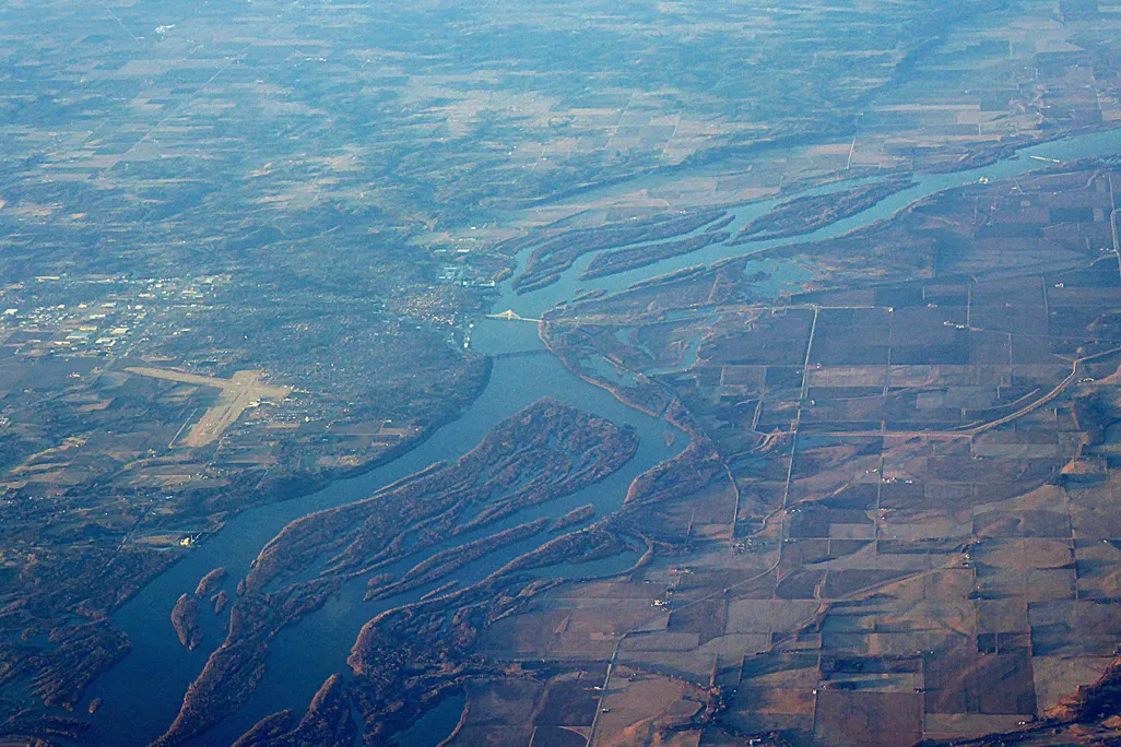 An aerial photo of the Mississippi River flowing through Iowa. The river runs through the middle of the photo, separating patches of fields on the right and more urban and suburban areas on the left. 