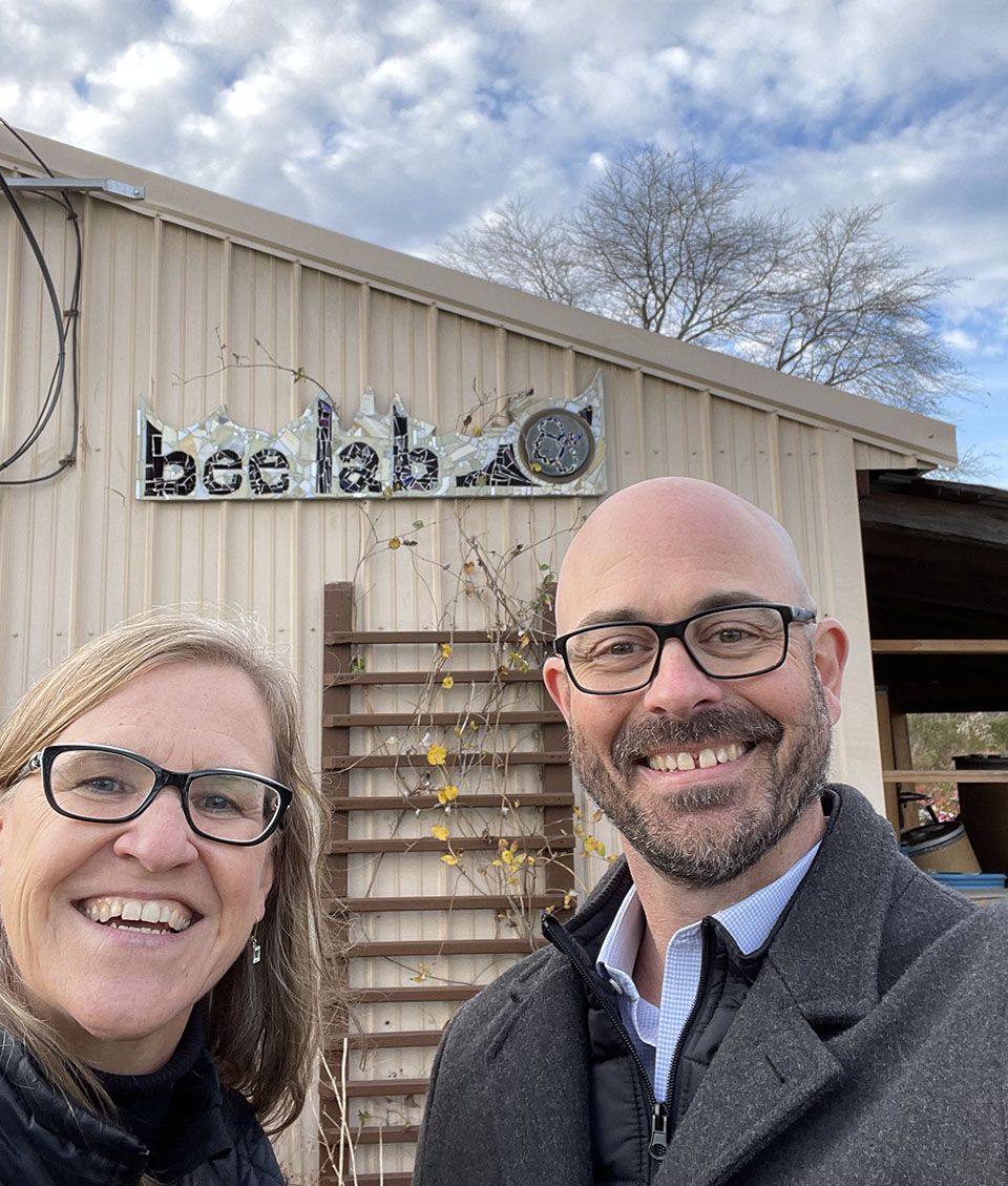 Two people pose in front of a beige barn with the sign Bee Lab on the building.