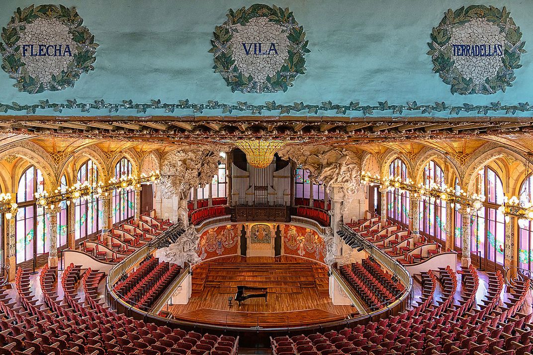 Interior of a music hall, with tiered seating arranged in a horseshoe shape. It is lit up by arched windows on either side.