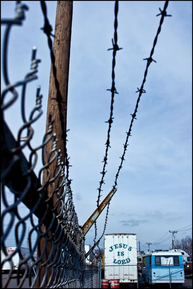 A truck with "Jesus is Lord” sits parked alongside a barbed-wire fence.