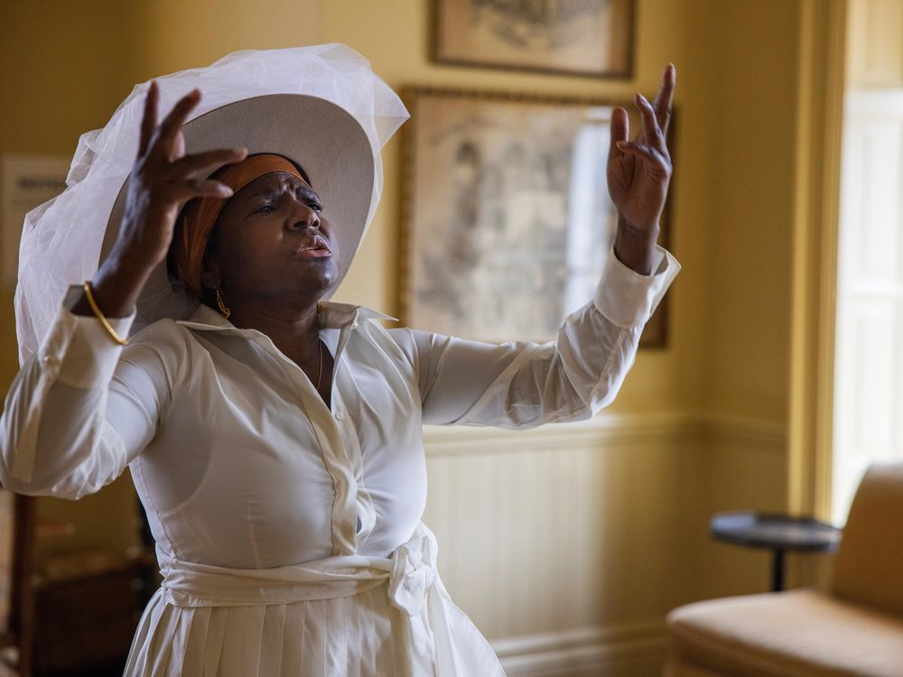 A black woman in a white dress and a large brimmed white hat stands in a room with yellow painted walls, raises her hands and closes her eyes, as though caught in the midst of dancing and singing