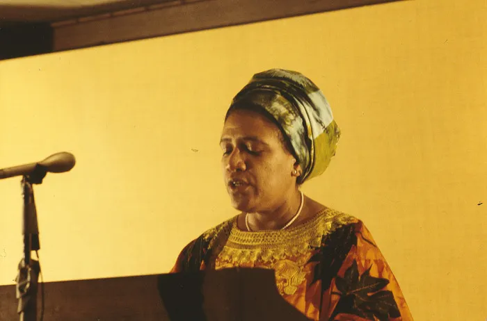 Candid color photo of Audre Lorde from the shoulders up standing at a microphone and podium.