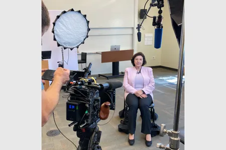 Annamarie Lusardi, seated in a chair facing a camera and lighting equipment.