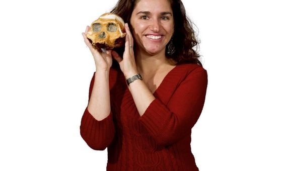 Headshot of paleoanthropologist Briana Pobiner in a red sweater holding an early human skull up text to her face on white background.