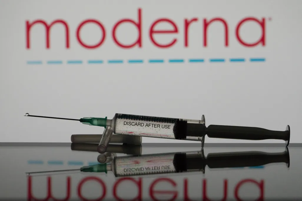 A vaccine needle lies in front of a Moderna logo