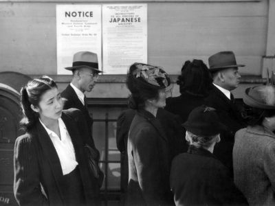 Japanese Americans stand in front of a poster with internment orders.