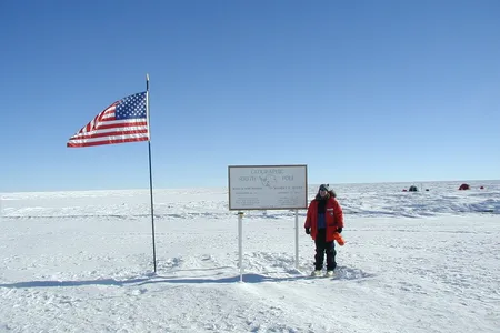 Geolgist Cari Corrigan poses for a picture in the snow next to an American flag and a sign that reads "Geographic South Pole."