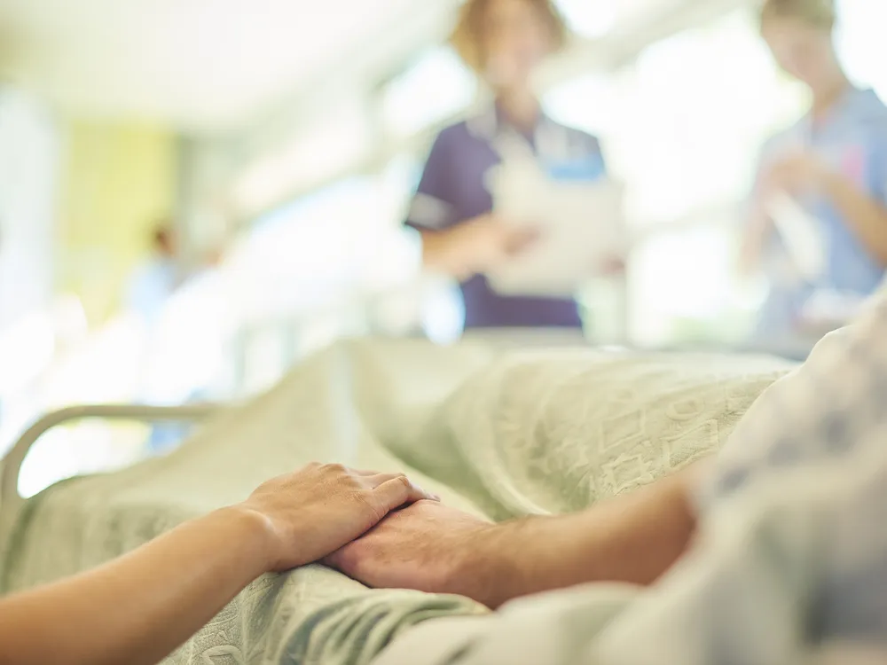 Close-up of person in hospital bed holding hands, with nurses blurred in the background