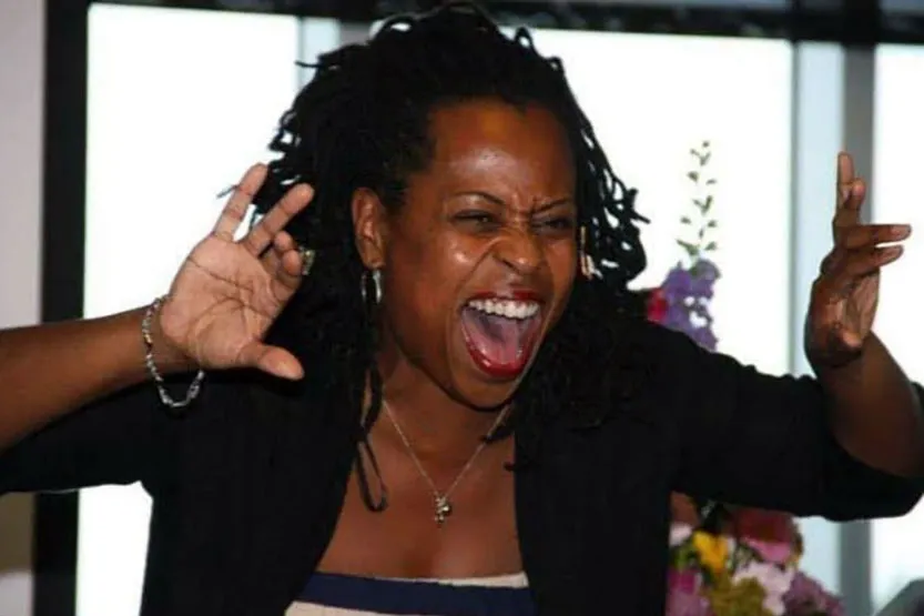 A Black woman acts out a part in a story, her hands raised as if pulling back curtains, her face menacing, like an animal about to attack.