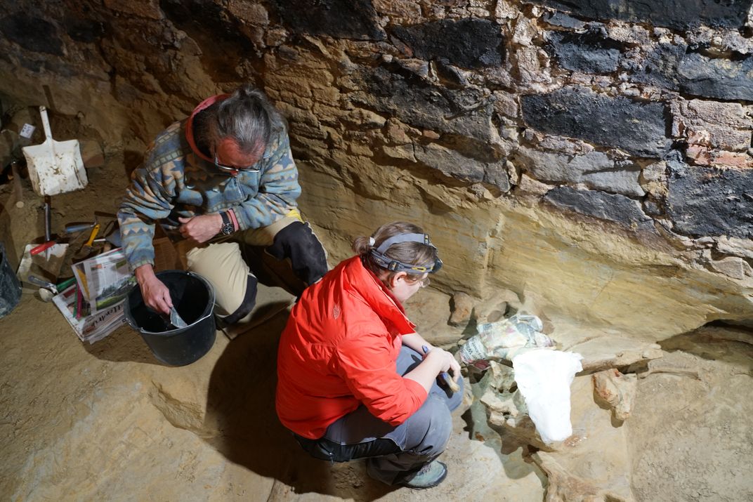 Woman in a red jacket brushing off a mammoth bone with a man kneeling nearby