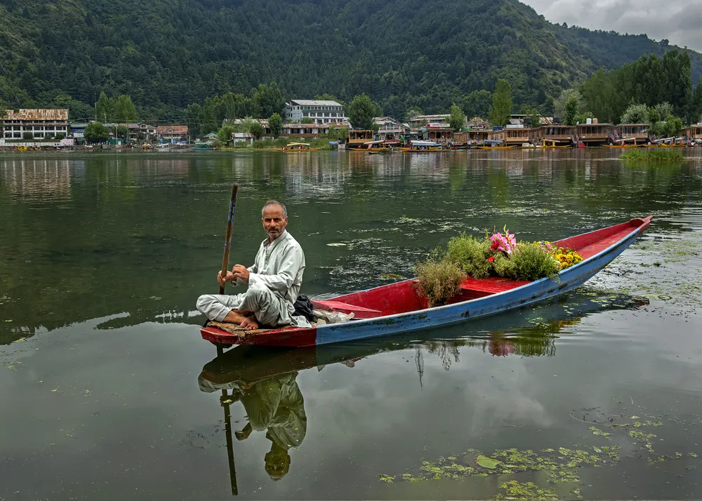 A man is selling flowers on his boat called shikara. He uses it to move around in Dal Lake of Kashmir and sell flowers to the tourists present in house boats.