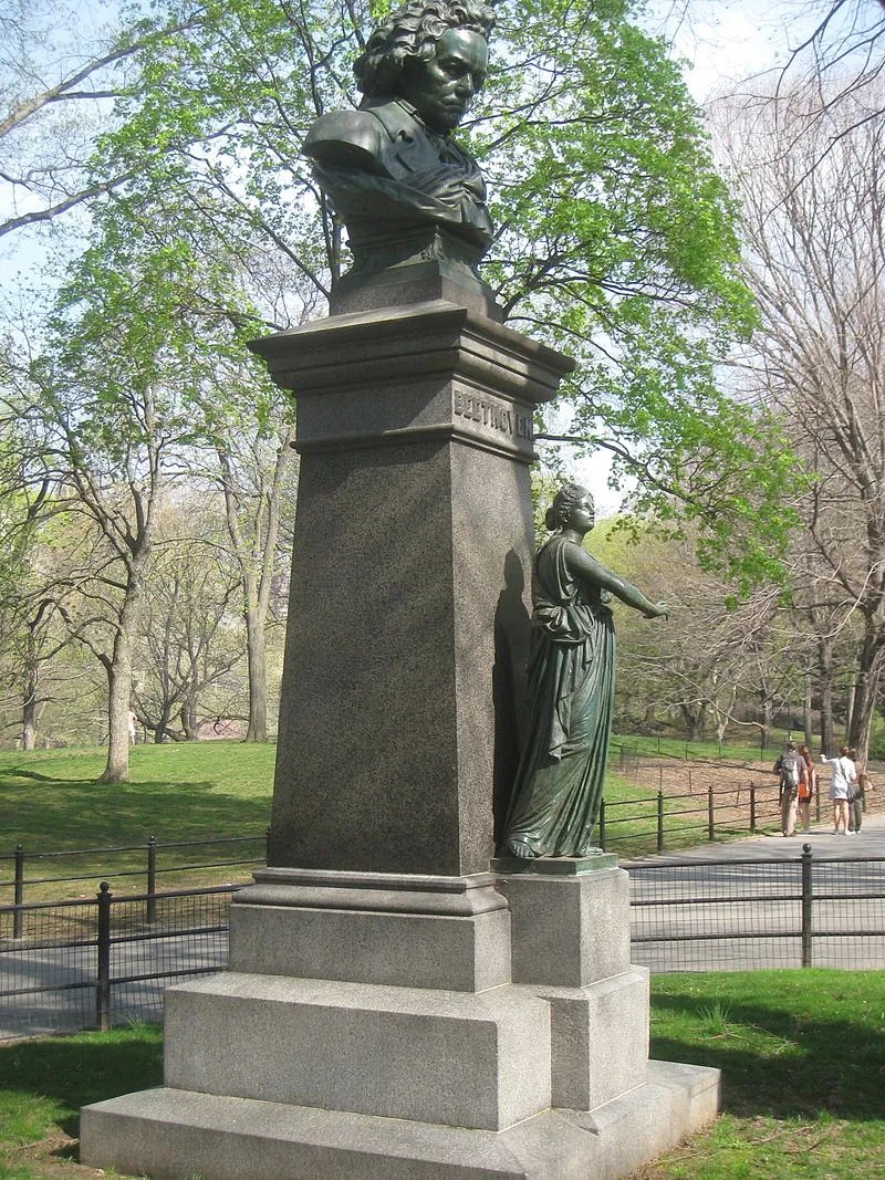 A pedestal surrounded by greenery in Central Park, with Beethoven's bust on top looking down and a smaller figure of a woman in robes standing beneath him