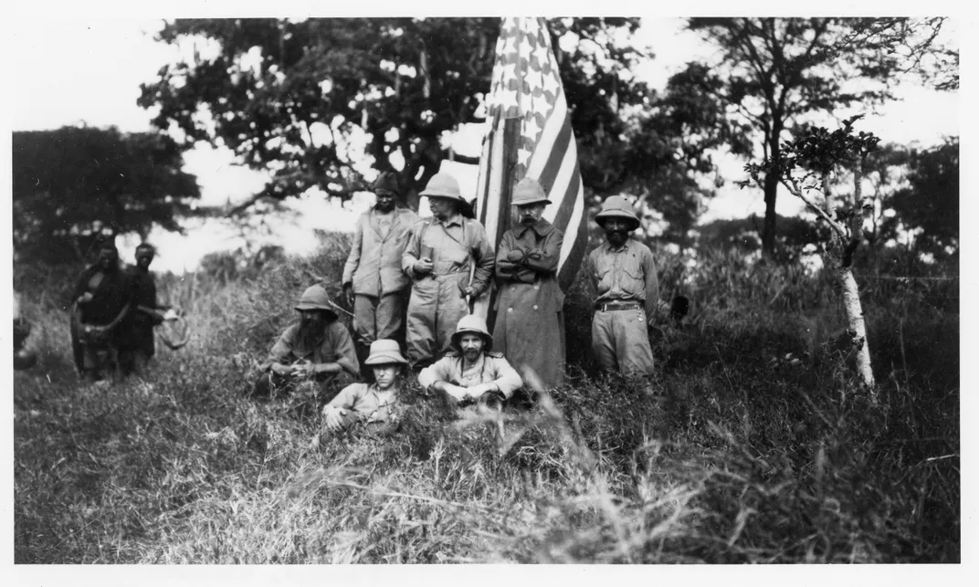 Members of the Smithsonian-Roosevelt African Expedition. Roosevelt is standing directly to the left of the flag, with his face turned away from the camera.