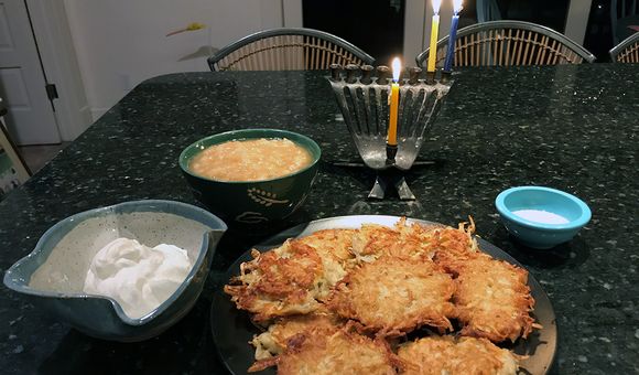A plate full of golden brown potato pancakes are placed on a dining table. Behind them are bowls of sour cream, apple sauce, salt, and a menorah with three candles lit.