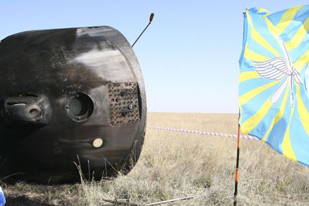 The Soyuz TMA-11 capsule lies on its side on the Kazakh steppe after landing. Search teams and companies involved in the rescue have planted their flags nearby.