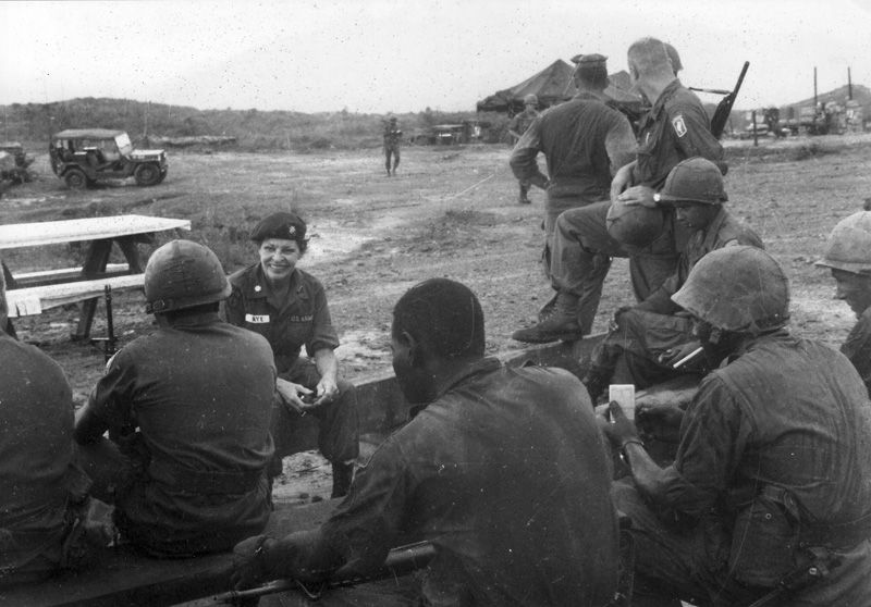 Martha Raye in conversation with members of the 173rd Airborne Brigade. Raye and the soldiers sit outside on benches. Tents, a jeep, and other soldiers can be seen in the distance in the background.