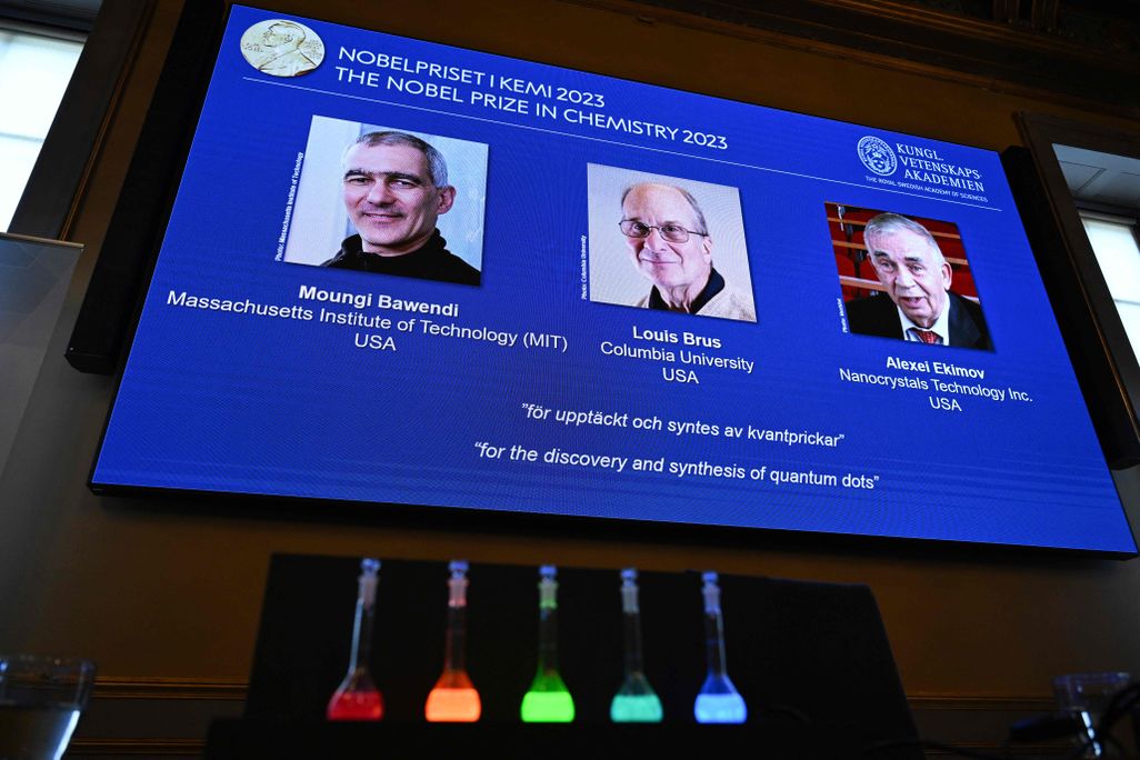 the winners of the chemistry nobel prize, Moungi Bawendi, Louis Brus and Alexei Ekimov, appear on a tv screen with five vials of glowing colors in front