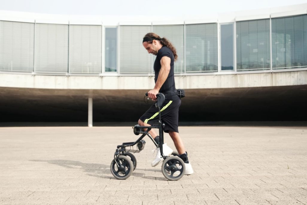 A man with long hair walks with the help of a walker on a concrete surface