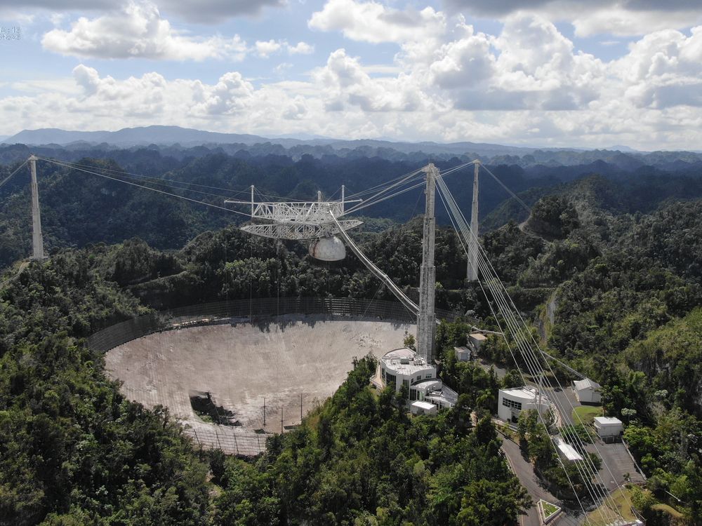 A photograph of the dish from high above. It has three tall towers around the circular dish, all connected by thick cables. The dish has a 100-foot hole on the side. The observatory is surrounded by expansive forest. 