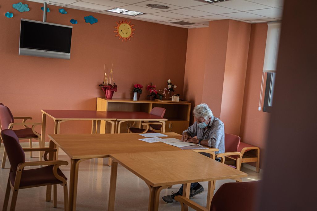 A person sits at a table wearing a mask and writing. The room in the nursing home is otherwise empty and has coral-colored walls