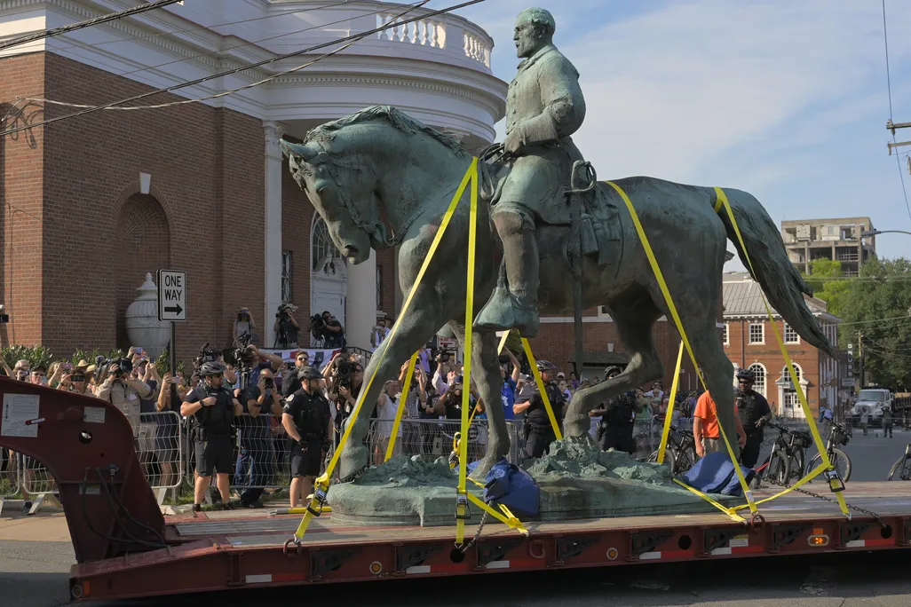 Construction crews transport a greenish bronze statue of Lee on horseback past a crowd of onlookers on a truck bed
