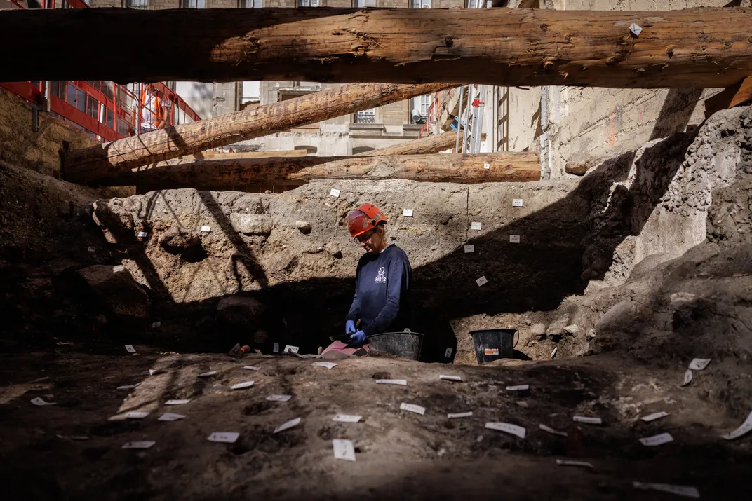 An archaeologist kneels in a deep trench, under wooden beams
