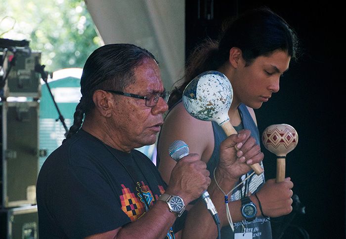 Two men with long hair, one older and one younger, perform with eyes closed on stage. They each hold a painted rattle, and the older man in the foreground also holds a microphone.