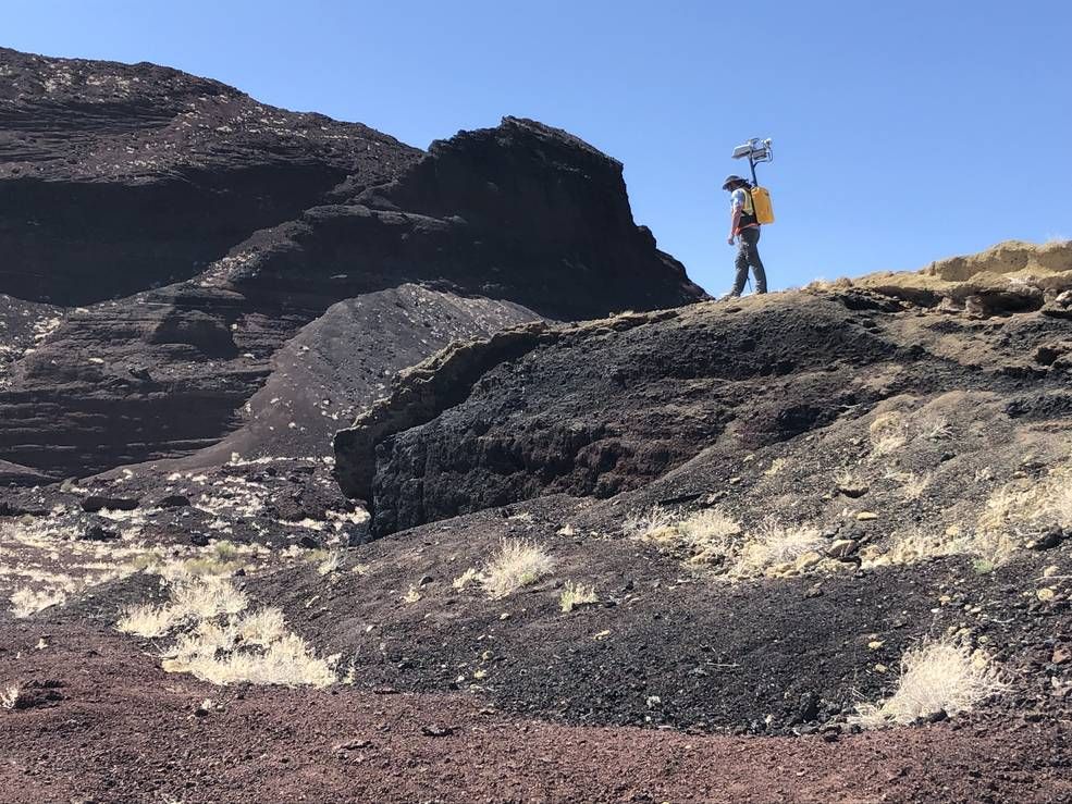 An image of a planetary scientist walking in a mountainous region with a yellow backpack on. The backpack has a camera attached to it.