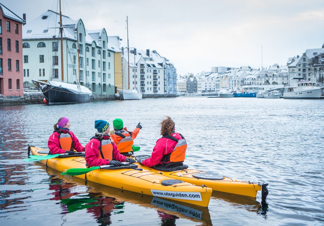 Four kayakers paddle through the harbor in Ålesund, Norway, surrounded by waterfront buildings, moored boats, and snow-dusted rooftops on an overcast day.