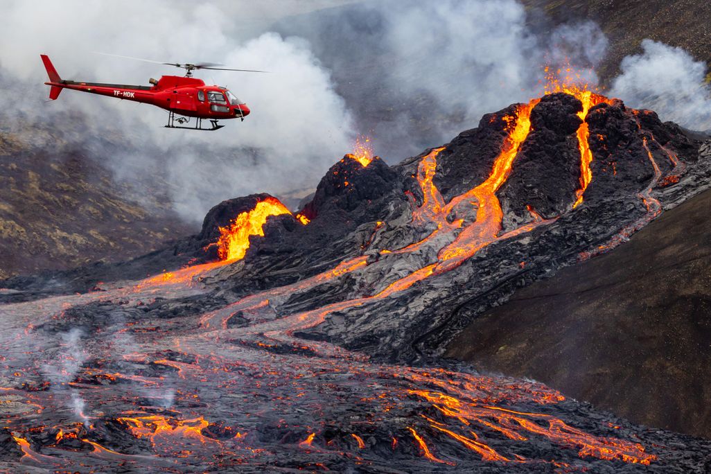 The photo shows a volcanic eruption. Magma is flowing down the volcano. A red helicopter hovers near the volcano.
