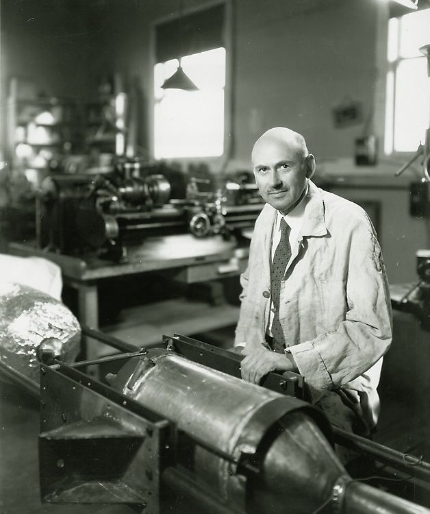 Black-and-white image of a white man in his early 50s. He is bald and has a mustache. Wearing a white lab coat, he is sitting at a work table supporting a large metal cylinder.