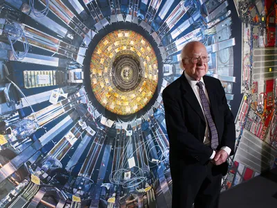 Peter Higgs stands in front of a photograph of the Large Hadron Collider at the Science Museum in London in 2013. The year before, researchers smashing protons together at the collidor had discovered evidence of a fundamental particle, which Higgs had proposed nearly 50 years prior.