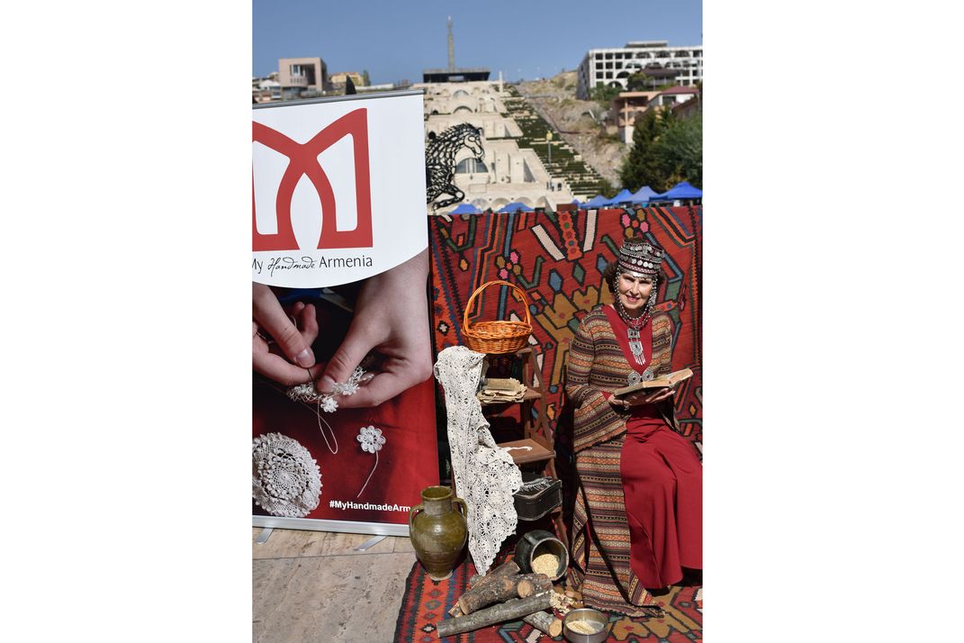 On a small, outdoor set covered in traditional rugs and textiles sits a woman. She wears traditional Armenian dress and hat.